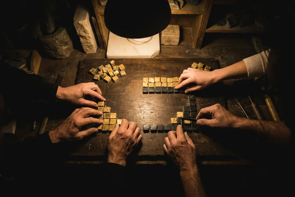 Manos de varias personas ordenando teselas doradas y oscuras en un taller